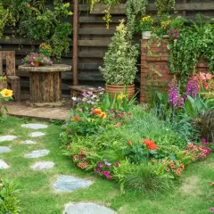 Ein üppiger, naturnaher Garten mit bunten Blumen, grünen Pflanzen und einem rustikalen Sitzbereich. Steinplatten führen durch das satte Grün.